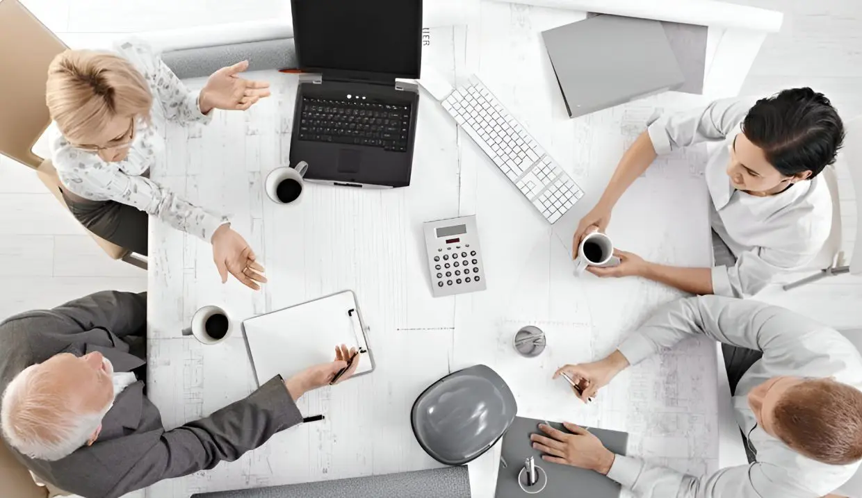 Four people sit around a table with laptops, papers, a calculator, and coffee cups, engaged in a business meeting or discussion.